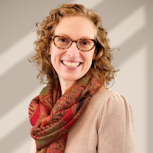 A smiling woman with curly hair wearing glasses, a beige sweater, and a patterned scarf against a softly lit tan wall.